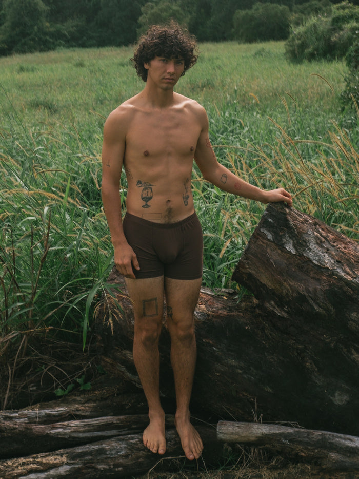 A shirtless man with curly hair stands outdoors in a grassy field next to a large log, wearing dark brown boxer briefs. He has visible tattoos on his chest and legs.