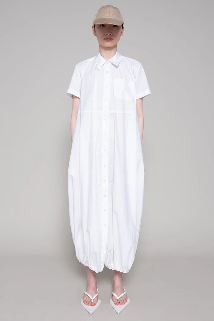 Person wearing a white dress with flip flops and beige cap on plain background