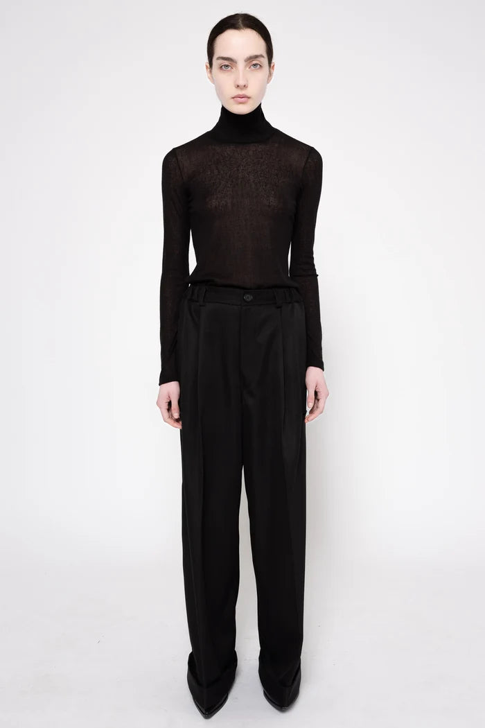 A model stands against a light gray studio background wearing a sheer black long-sleeve turtleneck top and high-waisted black wide-leg trousers.