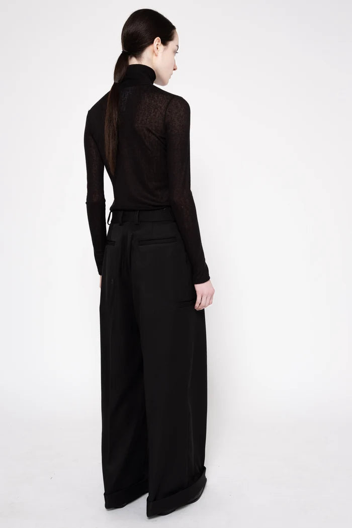 A model shown from the back wearing a black long-sleeve mock neck top with a sheer textured back panel and wide-leg black trousers, set against a plain white studio background.