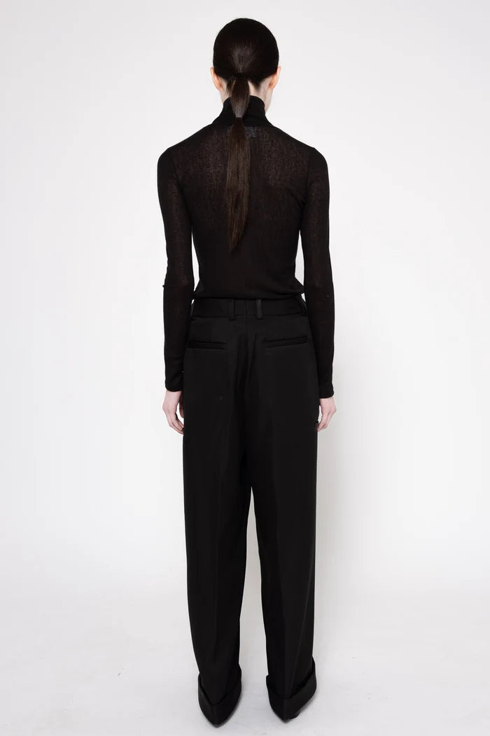 A model shown from the back wearing a black long-sleeve mock-neck knit top with sheer lace detailing and black wide-leg trousers, set against a plain white studio background.