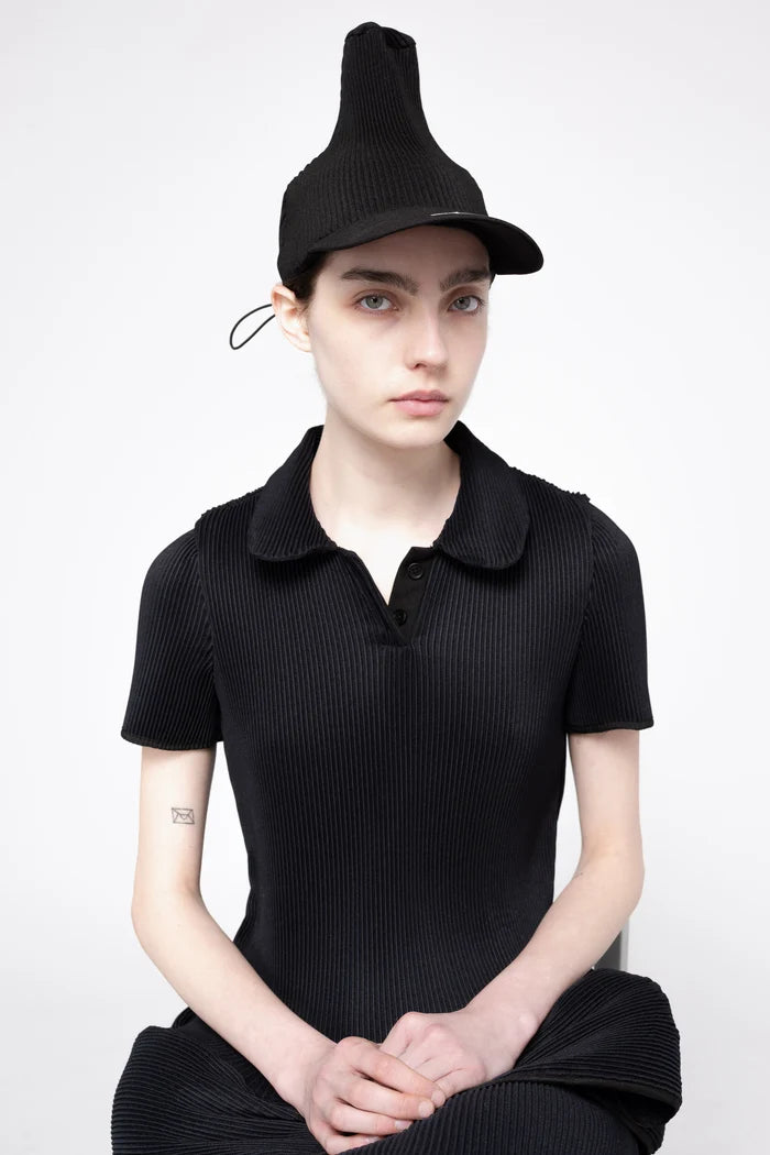 A model wearing a black pleated short-sleeve polo shirt, matching black pleated skirt, and a black pleated hat, posed against a light gray studio background.