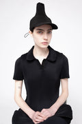 A model wearing a black pleated short-sleeve polo shirt, matching black pleated skirt, and a black pleated hat, posed against a light gray studio background.