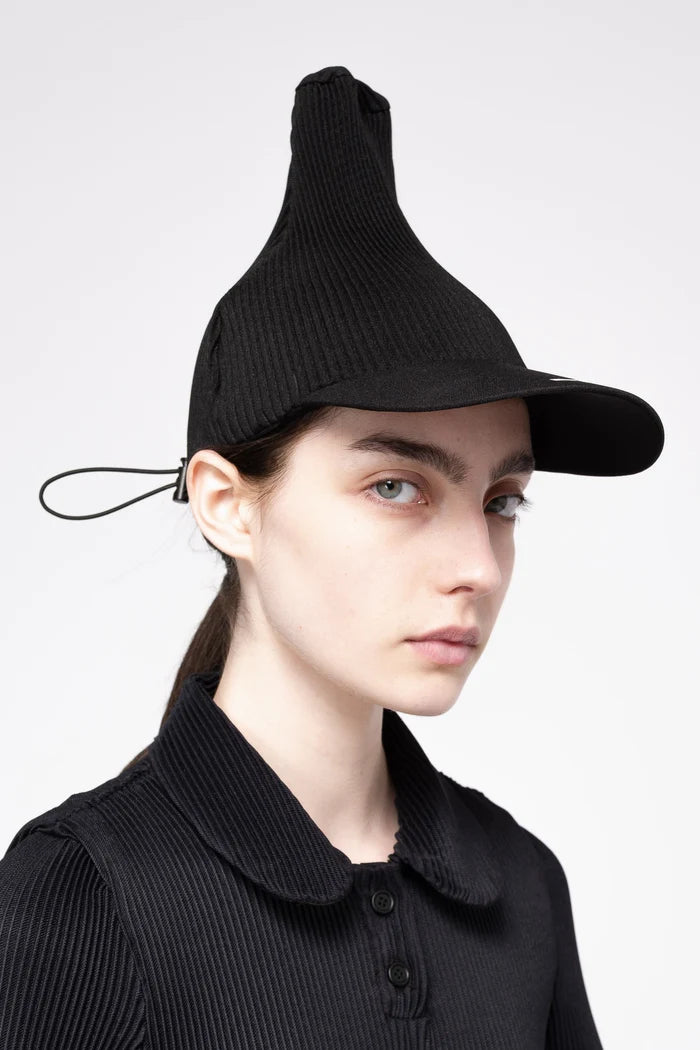 A model wearing a black pleated cap with a unique pointed top and short brim, paired with a matching black ribbed knit polo shirt, against a light gray background.