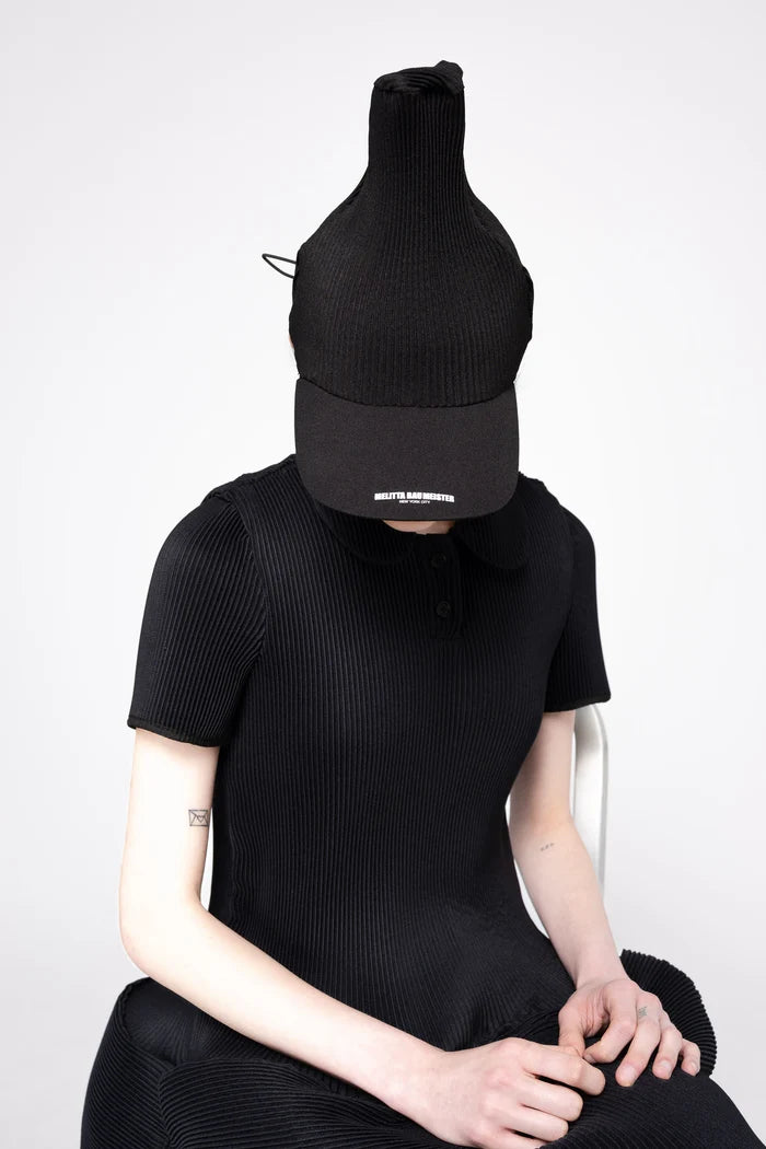 A person wearing a black pleated cap with a small white logo and a matching black short-sleeve ribbed top, seated against a light gray studio background.