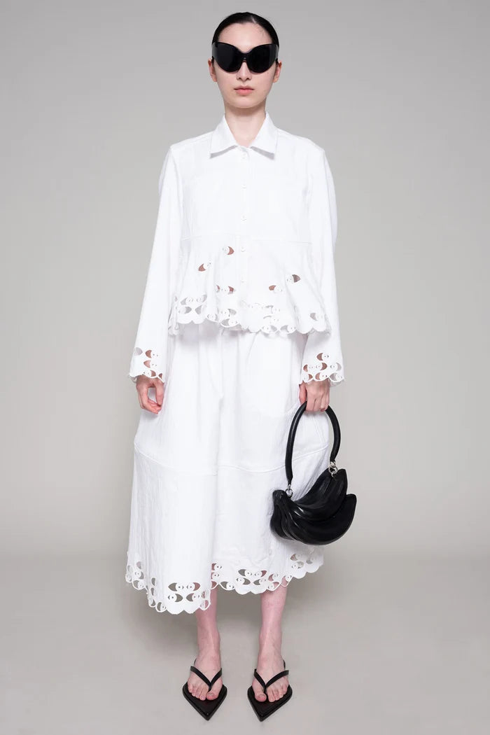 Person with black sunglasses wearing a white shirt and skirt with cut-out details holding a black bag on a plain background