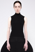 A model wearing a black sleeveless mock-neck top and a matching black skirt, photographed against a light gray studio background.