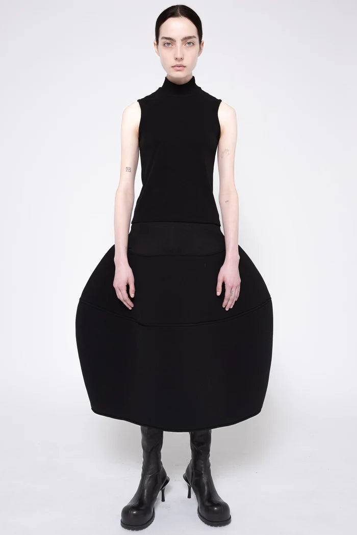 A model wearing a black sleeveless mock-neck dress with a very large, structured balloon skirt and black heeled boots, photographed against a light gray studio background.
