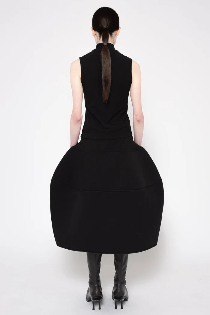 A model shown from the back wearing a black sleeveless dress with a high neck and a very large, voluminous skirt, paired with black knee-high boots against a plain white studio background.