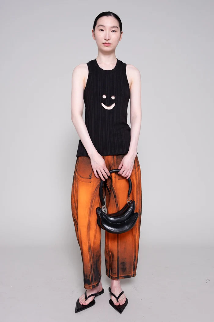 Person wearing a black sleeveless top with a smiley face design and orange pants, holding a black handbag on a gray background.
