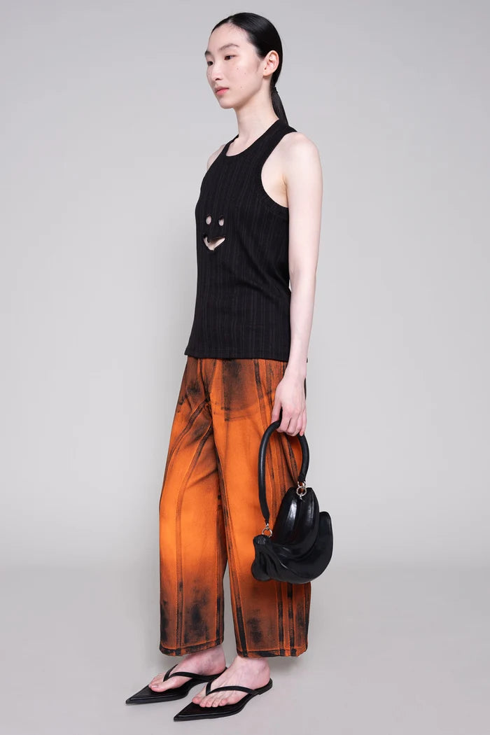 Person wearing a black sleeveless top and orange painted pants with black bag on a plain background