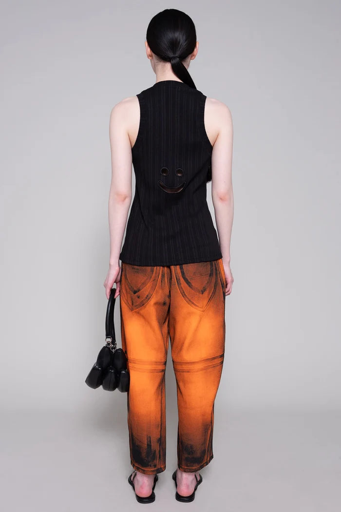 Person wearing a black sleeveless top and orange painted pants with black bag on a plain background