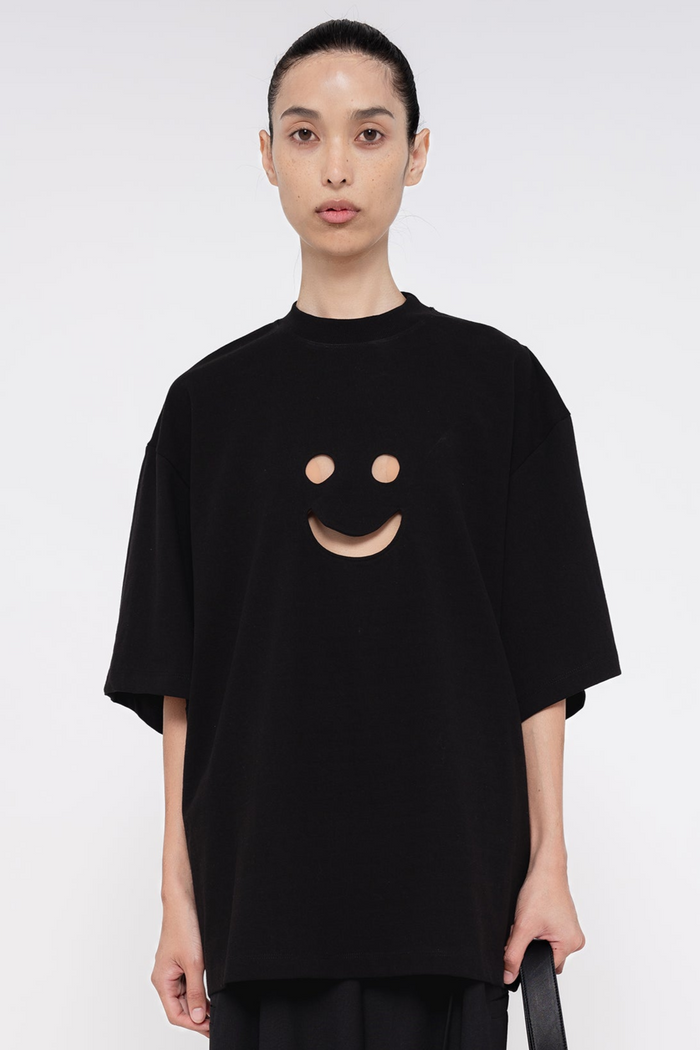PERSON-WEARING-BLACK-TEE-WITH-SMILEY-FACE-CUT-OUT-AND-BLACK-PANTS-HOLDING-BLACK-BAG-CLOSE-UP-FRONT-VIEW
