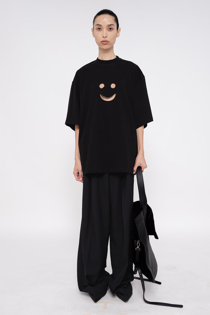 PERSON-WEARING-BLACK-TEE-WITH-SMILEY-FACE-CUT-OUT-AND-BLACK-TAILORED-PANTS-HOLDING-BLACK-SLOUCHY-WEEKENDER-BAG-WITH-POINTY-BLACK-BOOTS-FRONT-VIEW