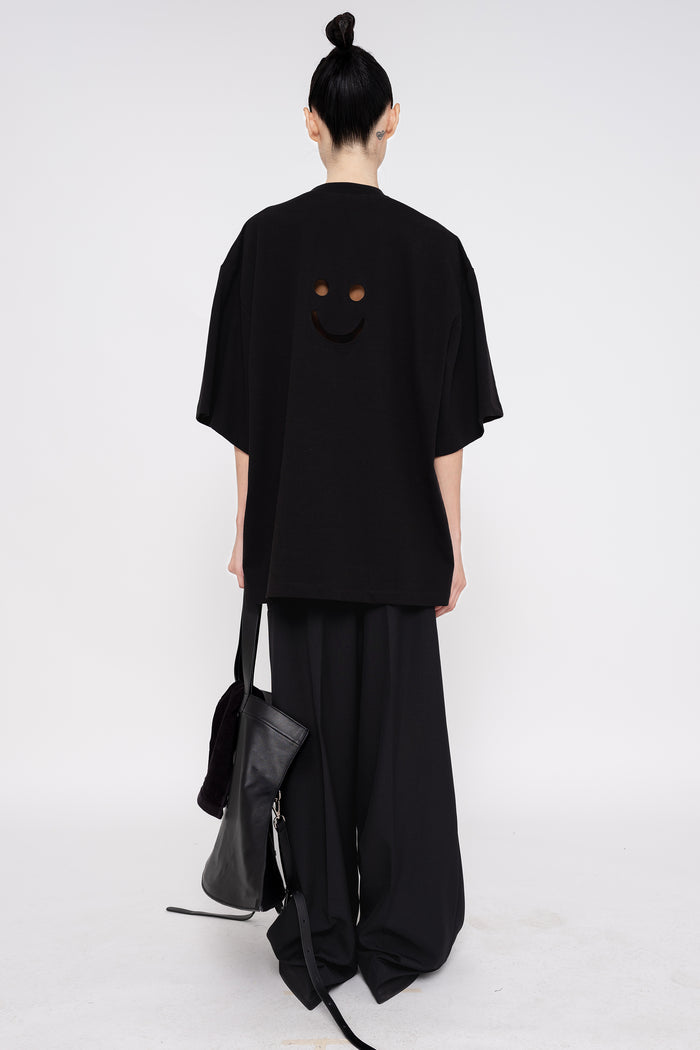 PERSON-WEARING-BLACK-TEE-WITH-SMILEY-FACE-CUT-OUT-AND-BLACK-TAILORED-PANTS-HOLDING-BLACK-SLOUCHY-WEEKENDER-BAG-WITH-POINTY-BLACK-BOOTS-BACK-VIEW