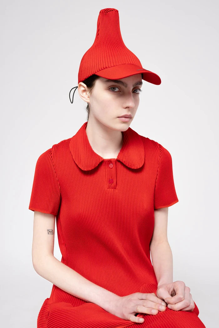 A model wearing a matching red pleated cap and short-sleeve polo dress, posed against a light gray studio background.