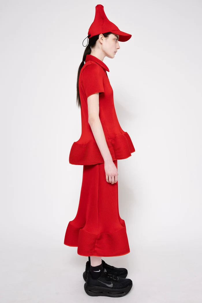 A model in profile wearing a bright red two-piece outfit with a short-sleeve collared top, matching wide-leg skirt, and a pointed red hat, paired with black sneakers, against a plain white studio background.