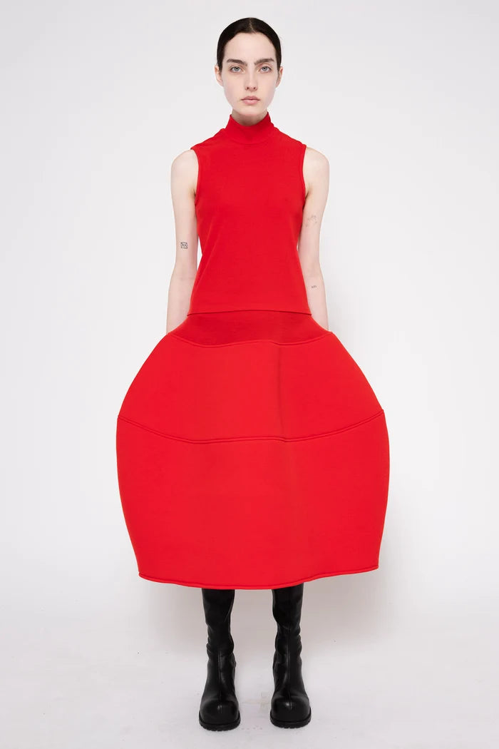 A model wearing a bright red sleeveless mock-neck dress with a very large, structured balloon skirt, paired with black boots, against a plain white studio background.