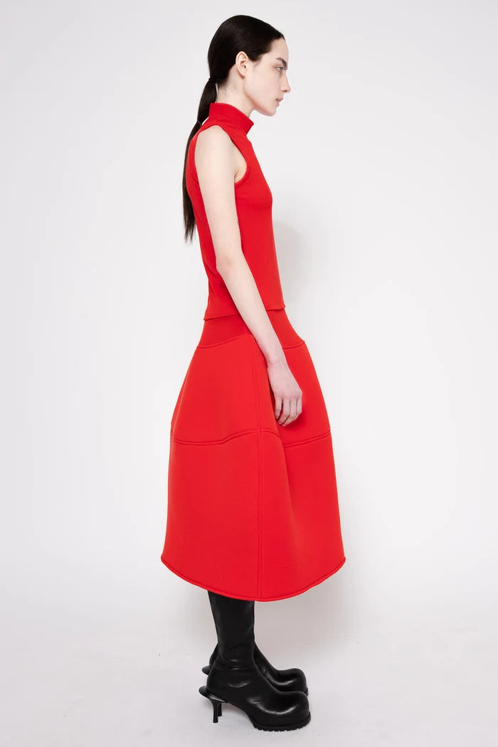 A woman shown from the side wearing a bright red sleeveless midi dress with a structured, balloon-like skirt and black knee-high boots, photographed against a light gray studio background.