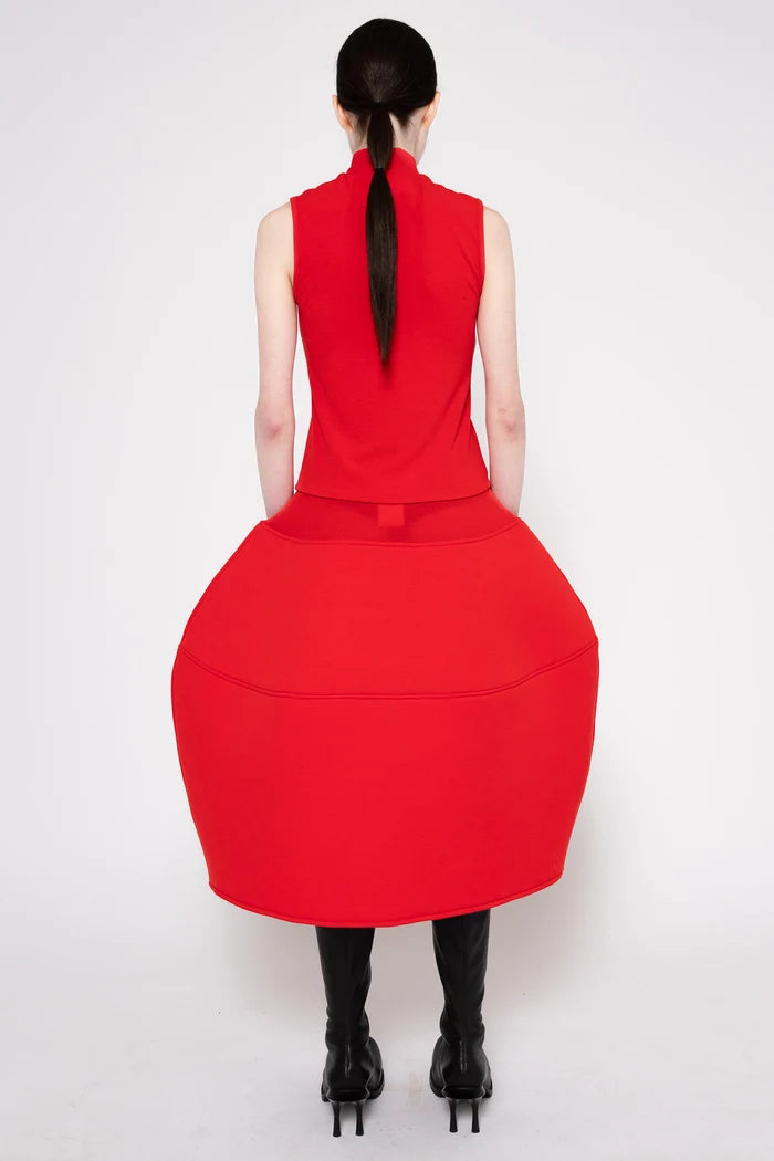 A model shown from the back wearing a bright red sleeveless midi dress with a high mock neck and a very full, balloon-like skirt, paired with black knee-high socks and black high-heeled boots, against a plain white studio background.