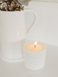 White Byron Bay candle in a glass holder  with a vase with dry flowers in the background