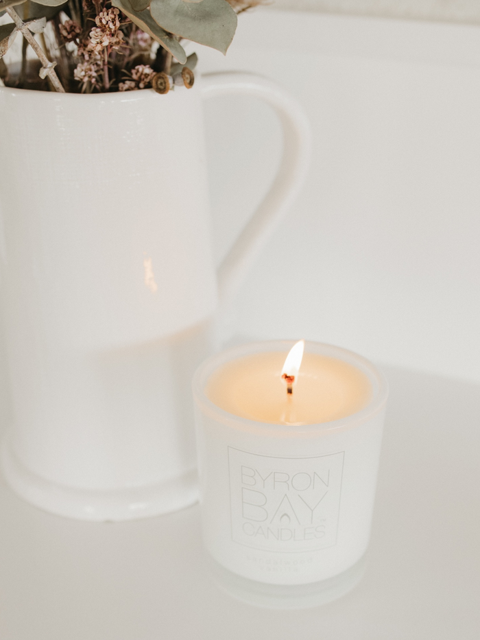 White Byron Bay candle in a glass holder  with a vase with dry flowers in the background