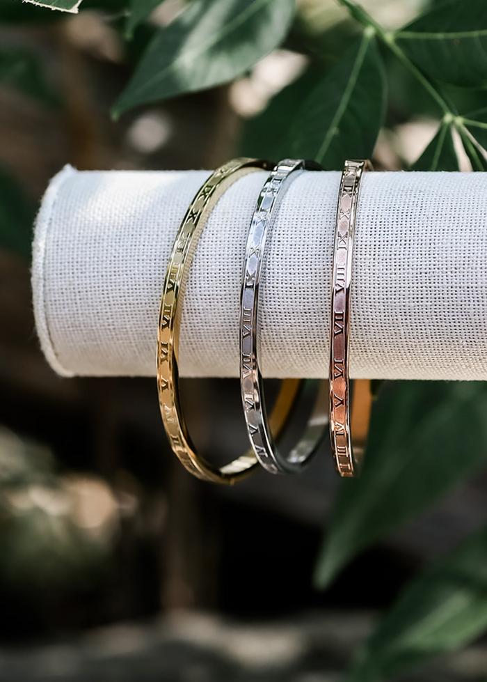 Three bangles with roman numeral etched detailing, displayed on a cylindrical object, with two gold-colored and one silver-colored bangle, set against a foliage background.