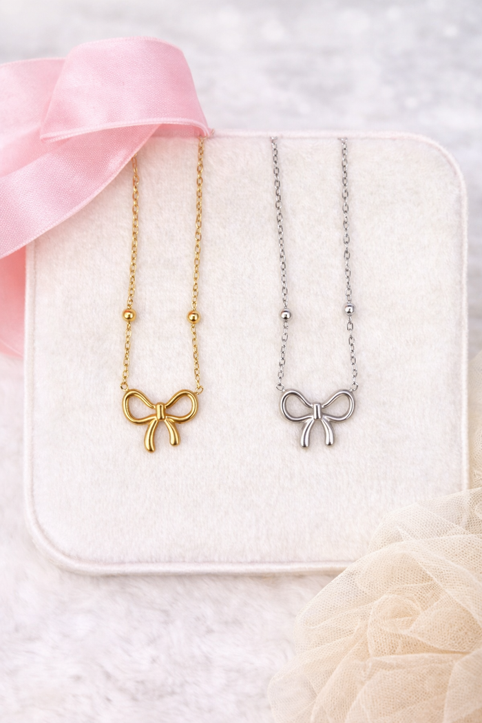 Two bow-shaped necklaces, one gold and one silver, on a white box with pink ribbon.