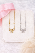 Two bow-shaped necklaces, one gold and one silver, on a white box with pink ribbon.