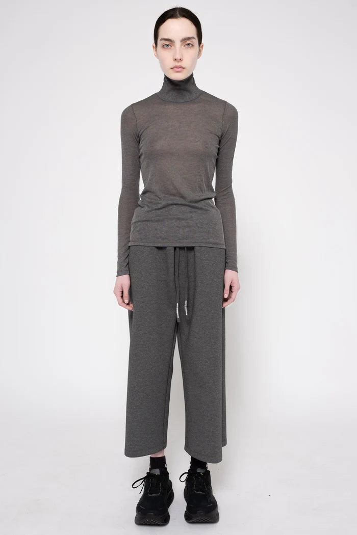 A model stands against a light gray studio background wearing a dark gray ribbed turtleneck sweater, matching wide-leg knit pants with a drawstring waist, black socks, and black sneakers.