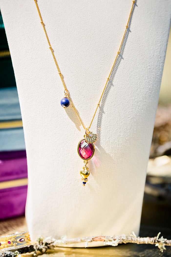Close-up of long gold layering necklace showing fan charm, lapis stone detail, and colorful glass pendant on delicate chain