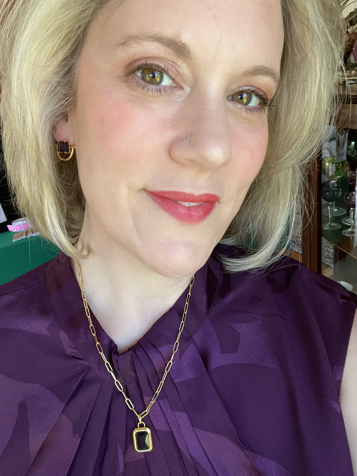 Woman wearing a purple top and gold necklace with a green gemstone.