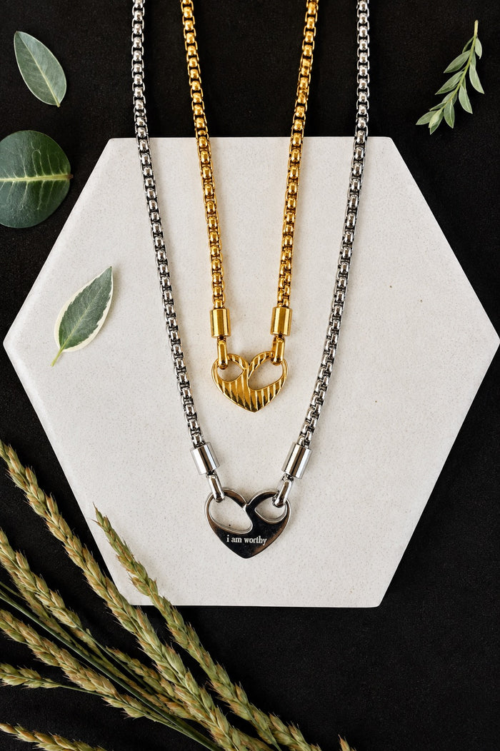 Two heart-themed necklaces displayed on a white hexagonal board against a black background, styled with dried wheat stalks and green leaves. One necklace is gold with a heart pendant, and the other is silver with a split heart pendant engraved with 'I am worthy'.
