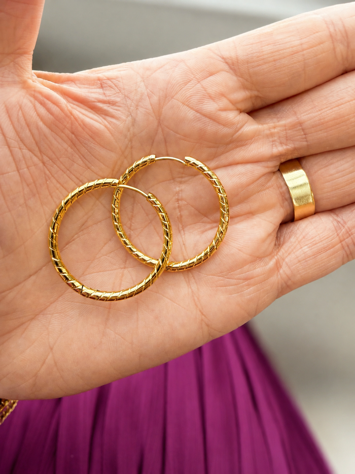 A pair of textured gold hoop earrings resting on an open hand, with a matching gold ring visible on the finger. The background is softly blurred, showing a gray wall and a purple garment.