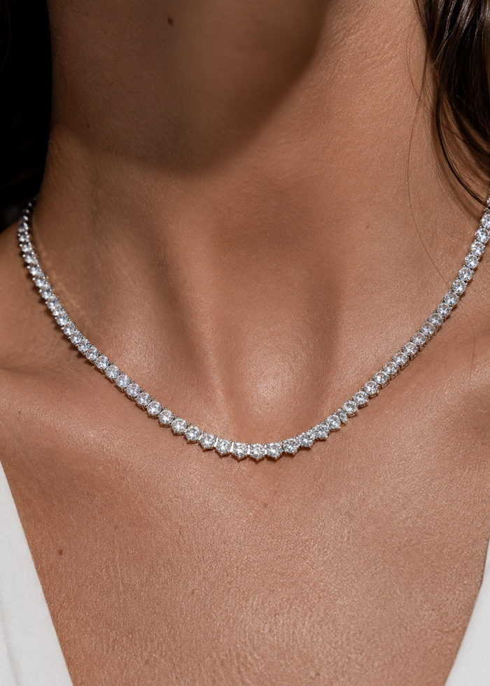 A close-up of a model's neck and upper chest wearing a delicate silver tennis necklace with clear stones.