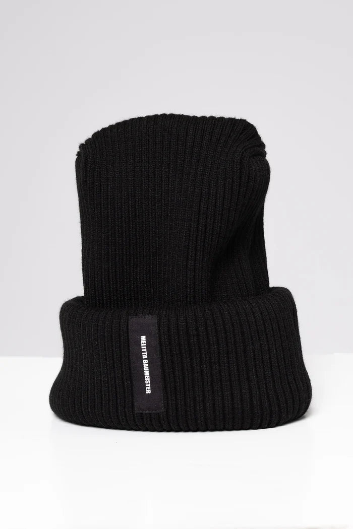 A black ribbed knit beanie hat with a folded cuff, featuring a small black vertical label on the side with white text that reads 'MELITTA BAUMEISTER'. The hat is photographed on a clean, light gray studio background.