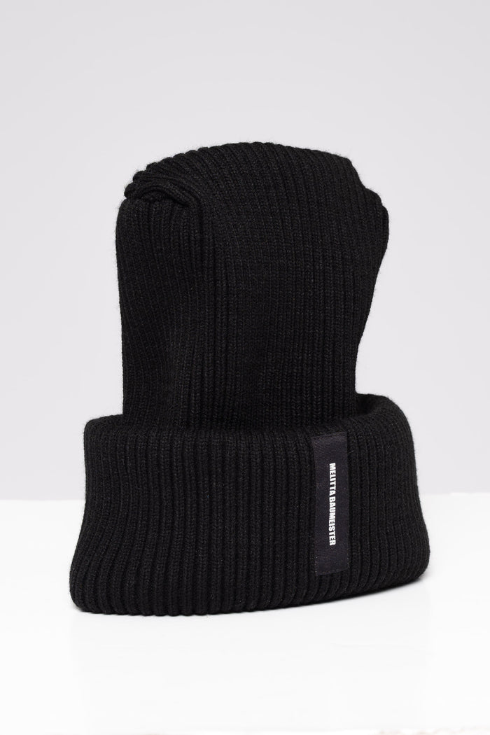 A black ribbed knit beanie hat with a folded cuff, featuring a small black vertical label on the side with white text that reads 'MELITTA BAUMEISTER'. The hat is photographed on a clean, light gray studio background.