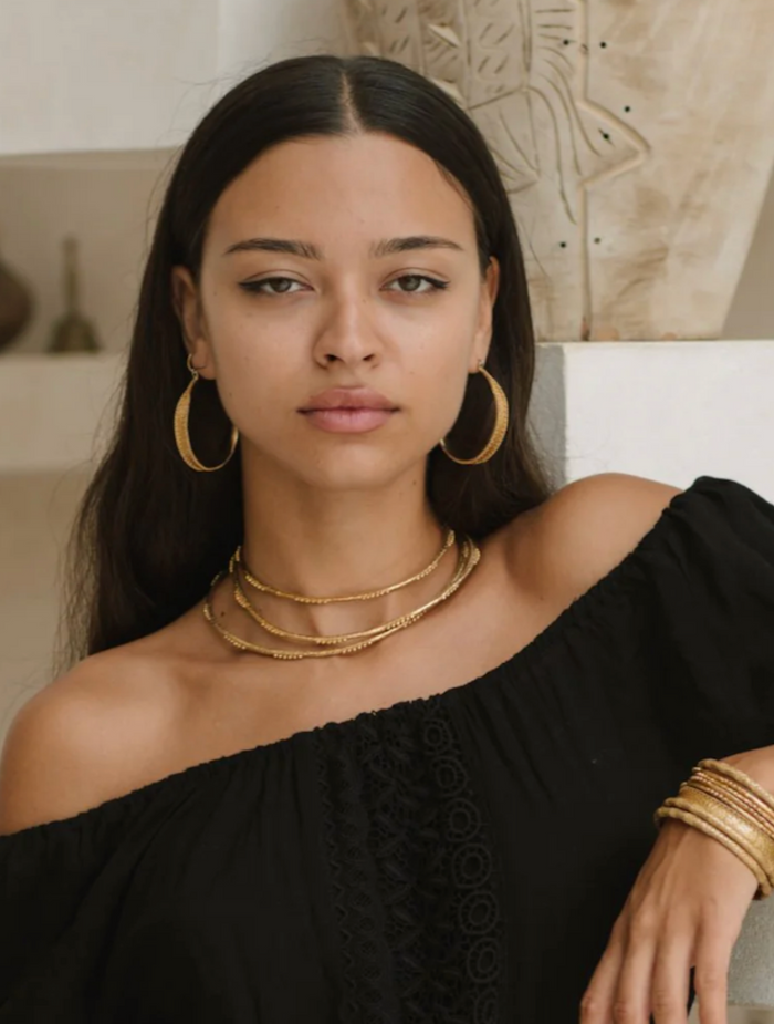 Woman wearing gold jewelry including earrings, necklaces, and bracelets in a neutral setting.