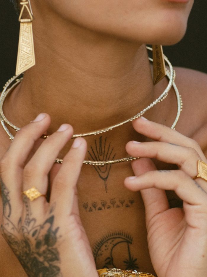 Close-up of a person wearing gold jewelry including earrings, necklace, and rings with tattoos on the neck and hands.