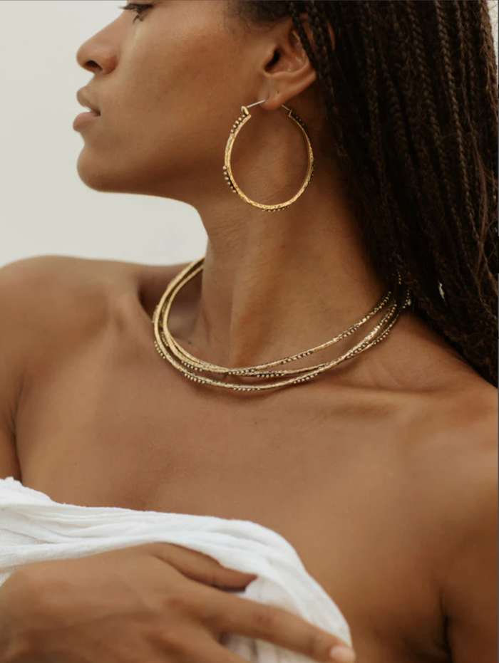 Woman wearing brass hoop earrings and matching chocker necklaces against a neutral background