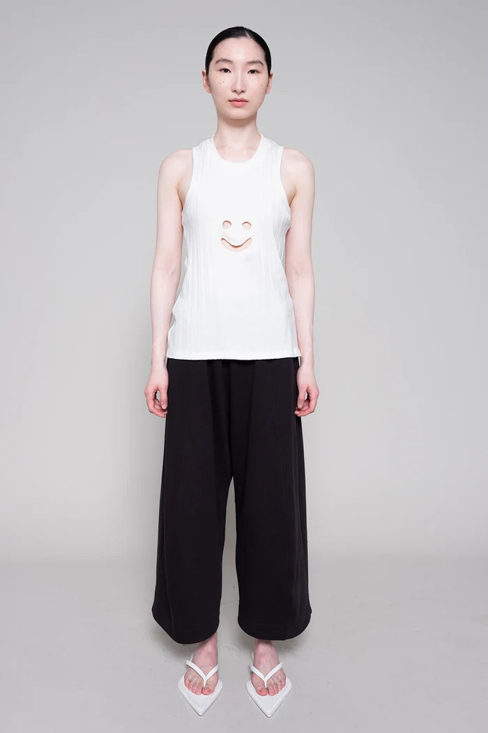 Person wearing a white sleeveless top with a smiley face design and black pants on a gray background