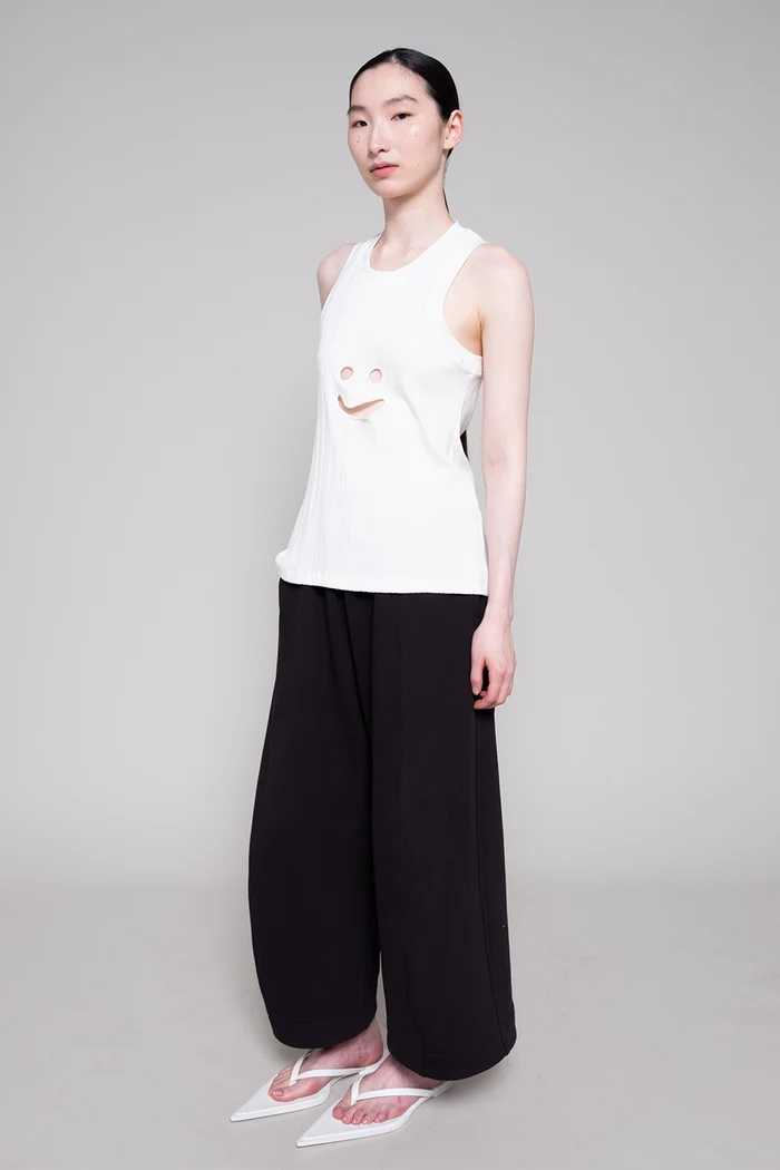 Person wearing a white sleeveless top with a smiley face design and black pants on a gray background